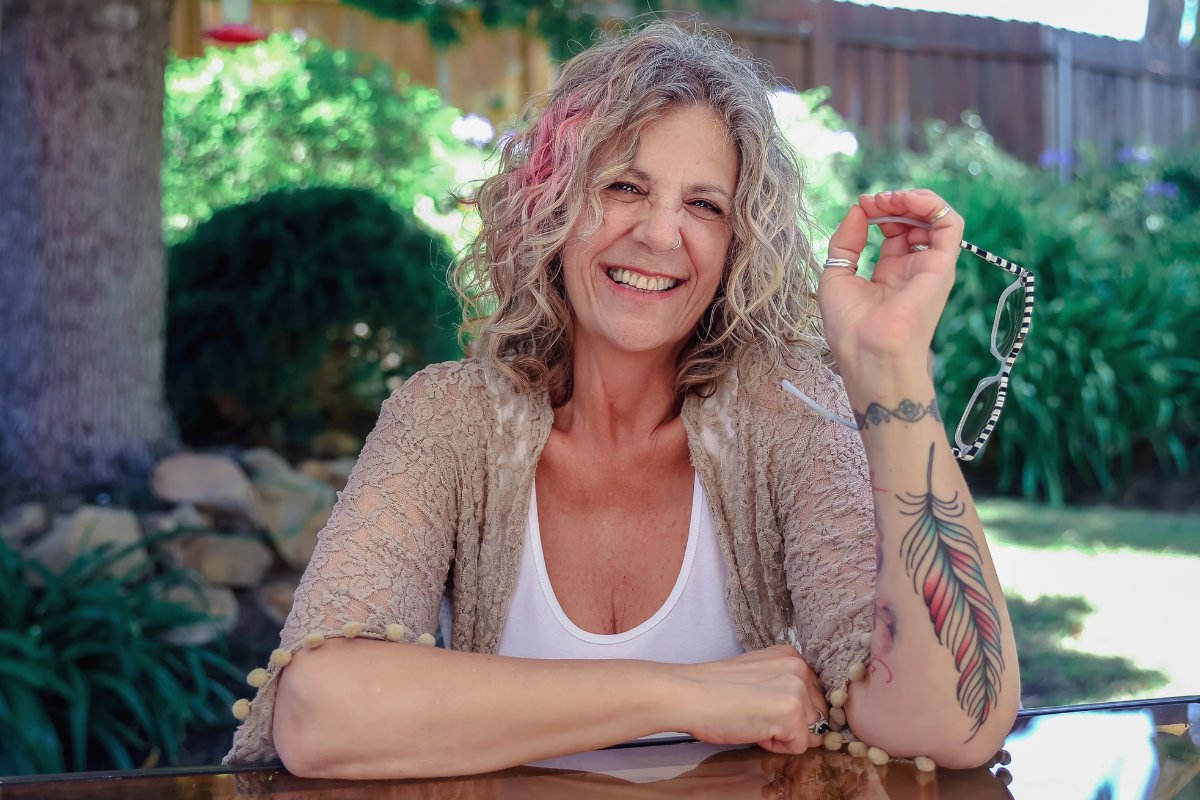 Femme mature souriante assise dans un jardin, tatouage de plume colorée fine sur l'avant-bras gauche, réalisé par Karine au studio Kanea à Toulon