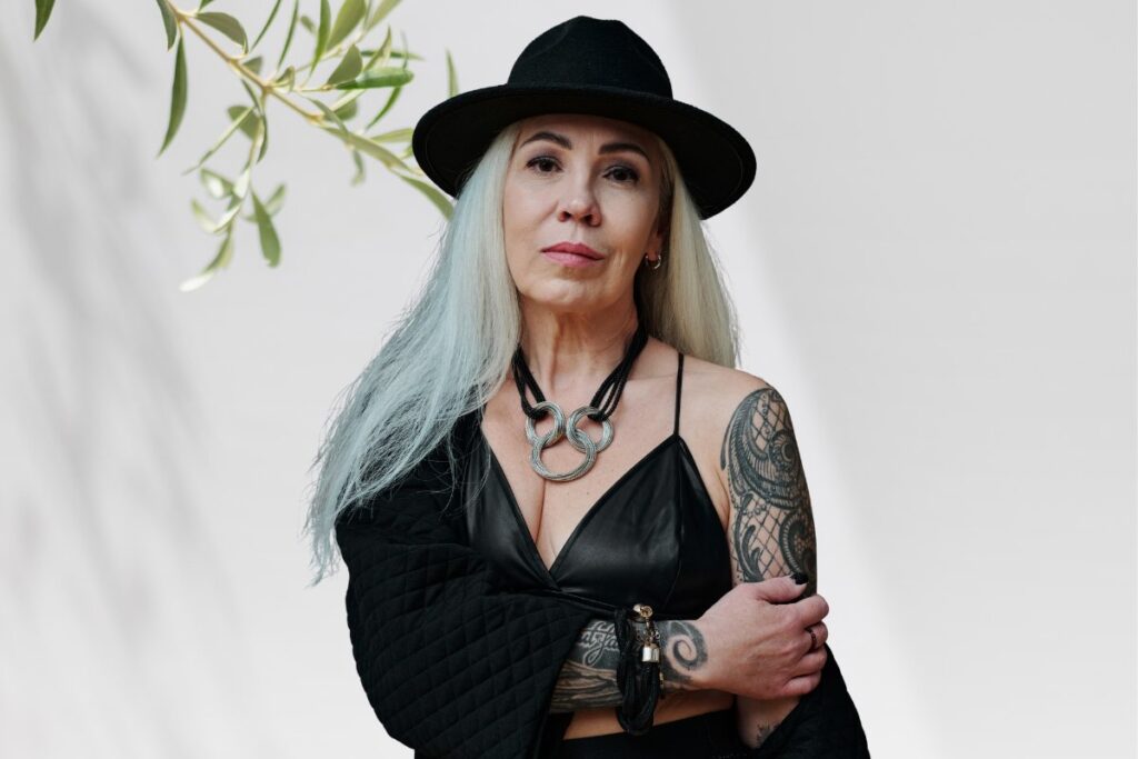 Femme mature aux longs cheveux gris-blonds avec chapeau noir, tatouage ornemental sur le bras, photographiée près d'une branche d'olivier au studio Kanea à Toulon