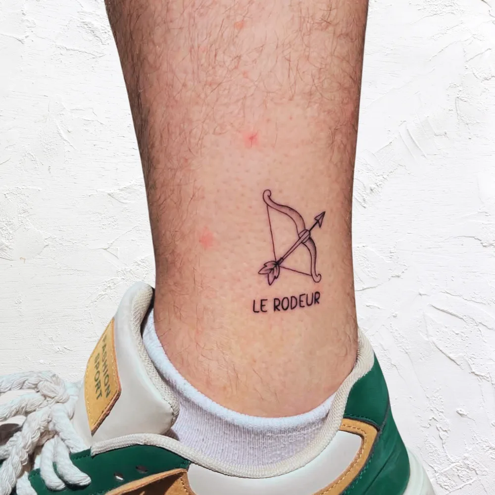 Tatouage minimaliste à la cheville : arc et flèche avec le mot Le Rôdeur, Kanea Toulon