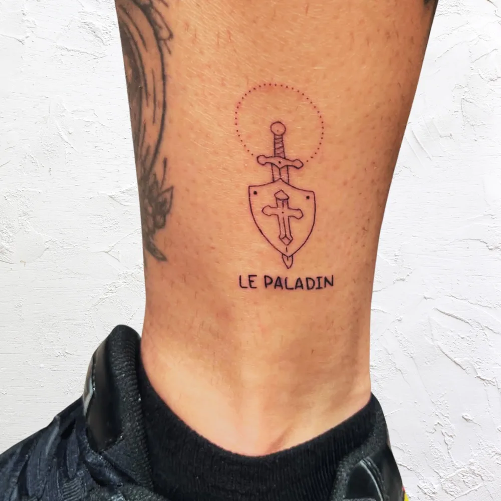 Tatouage fine line à la cheville : épée plantée dans un bouclier à croix, halo en pointillés et le mot Le Paladin, Kanea Toulon