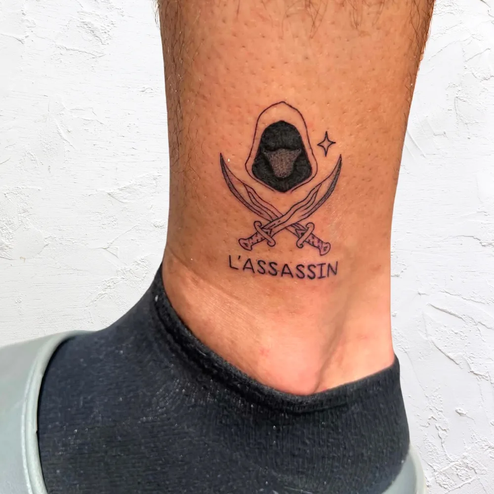 Tatouage fine line à la cheville : silhouette encapuchonnée et deux lames croisées avec le mot L'Assassin, Kanea Toulon